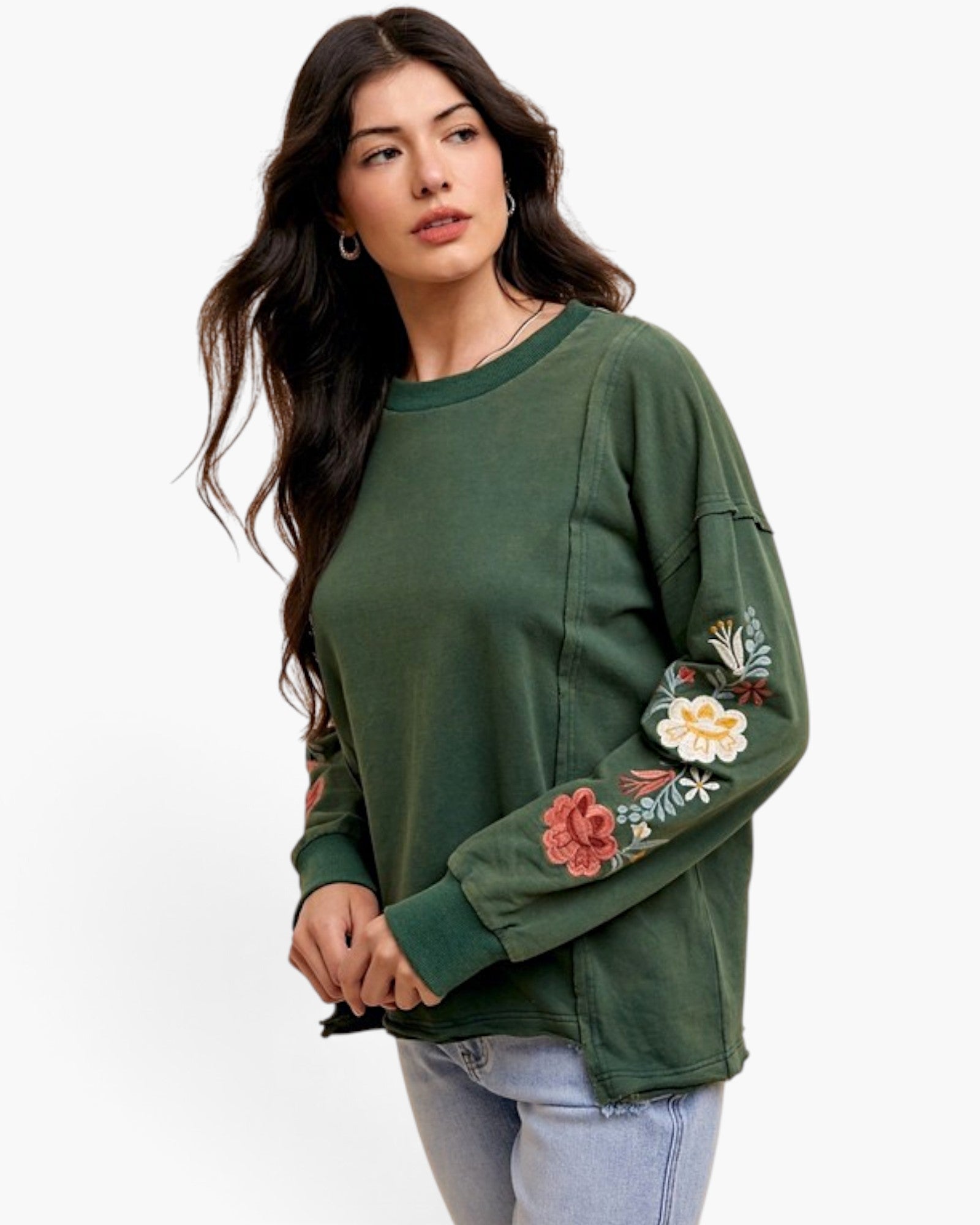 A woman wearing a dark green crewneck sweatshirt with colorful floral embroidery on the sleeves, paired with light blue jeans, against a plain white background.
