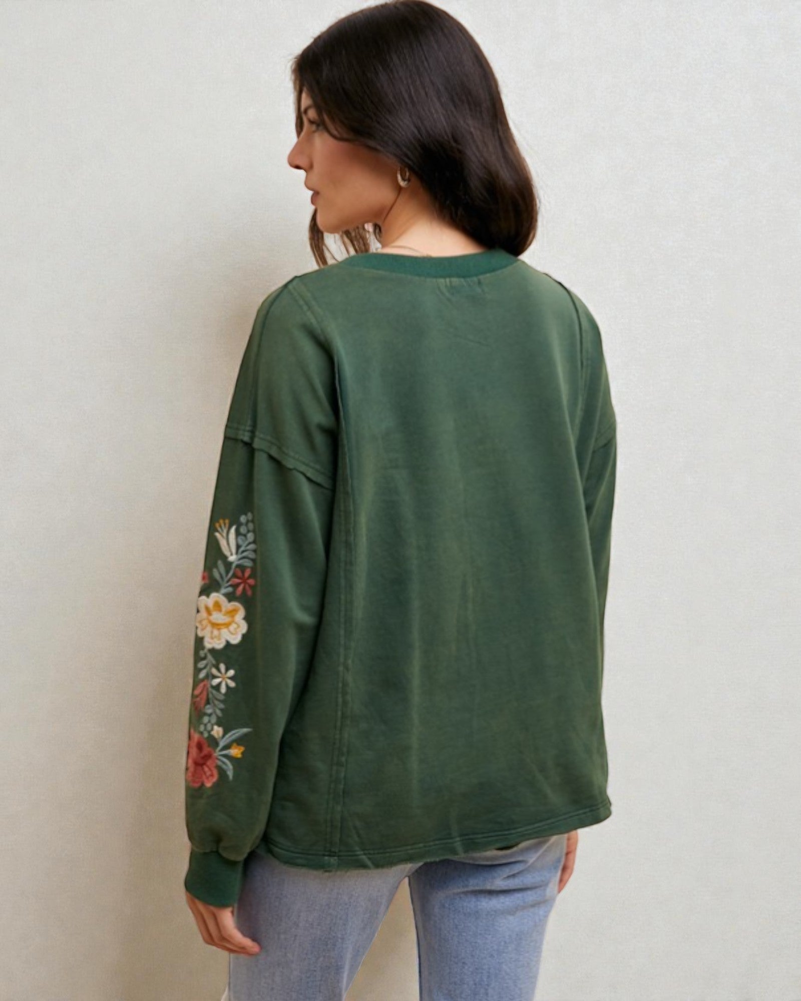 Woman wearing a green sweatshirt with floral embroidery on the sleeves against a plain background