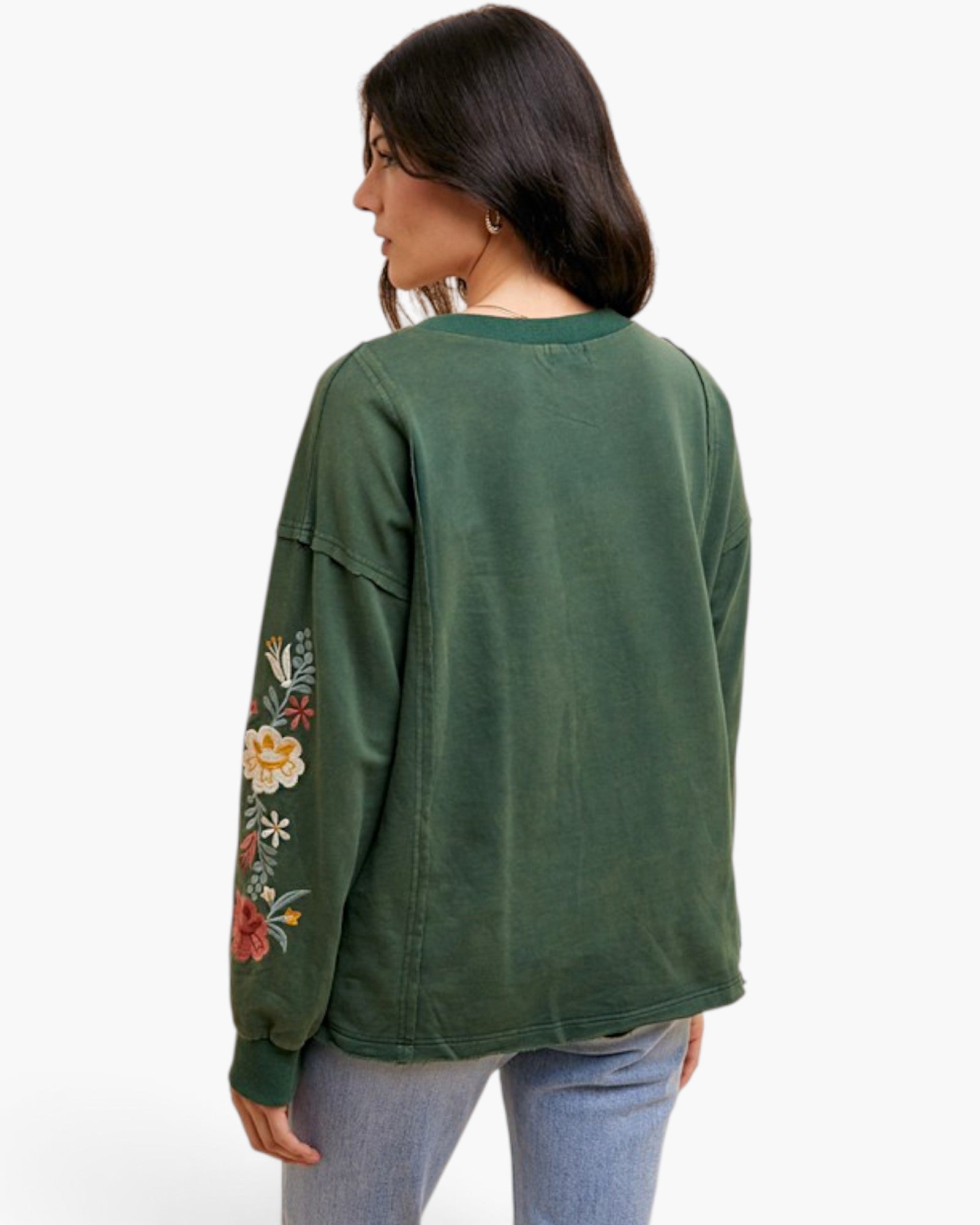 A woman shown from the back wearing a dark green long-sleeve sweatshirt with colorful floral embroidery on the left sleeve, paired with light blue jeans, against a plain white background.