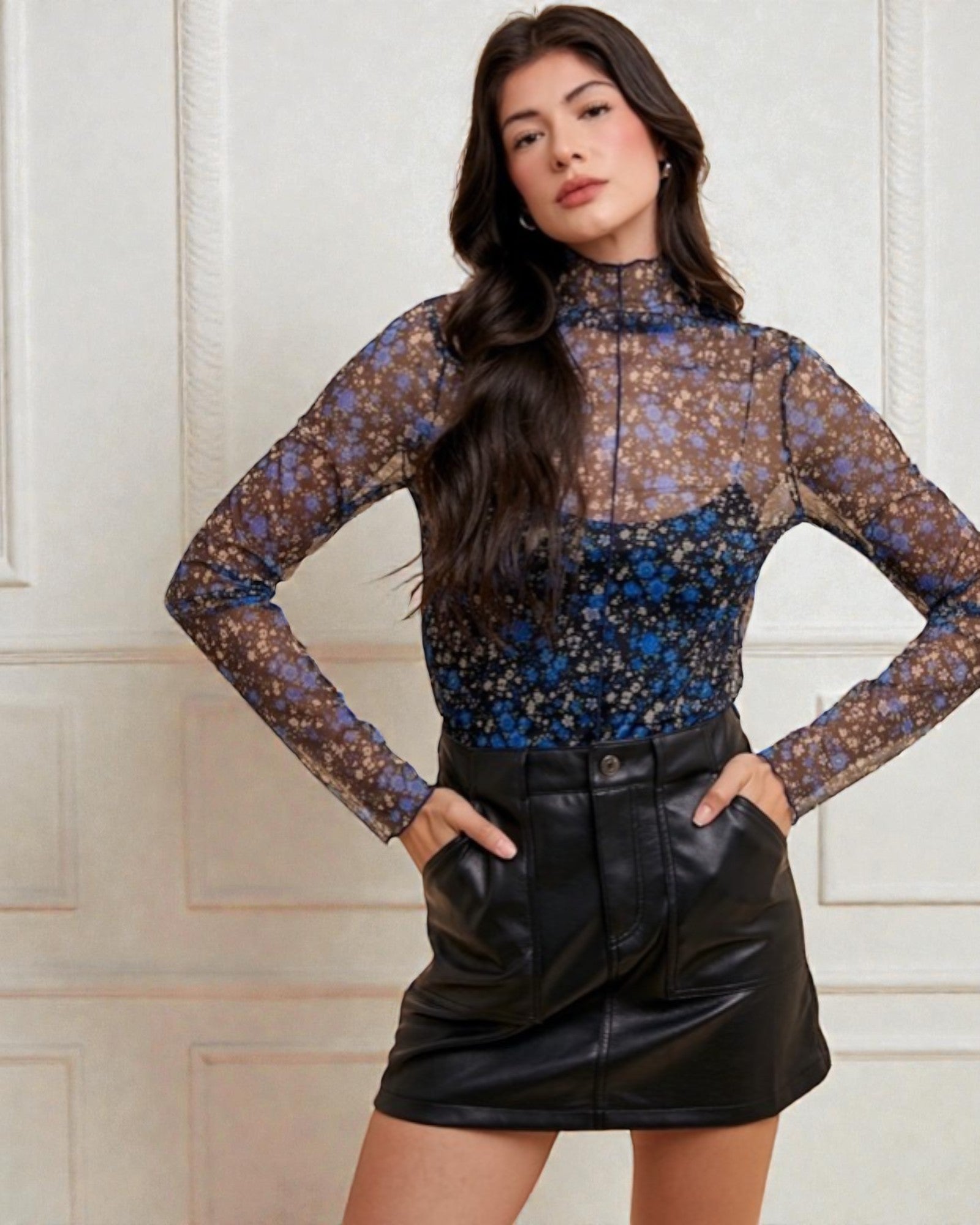 Woman wearing a floral long-sleeve top and black leather skirt against a white paneled wall.