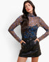 A woman modeling a sheer long-sleeve mock-neck top with a brown, blue, and gold floral pattern, paired with a black faux-leather mini skirt, against a plain white background.