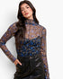 A woman modeling a sheer long-sleeve mock-neck top with a brown, blue, and beige floral pattern, paired with black faux-leather pants, against a plain white background.