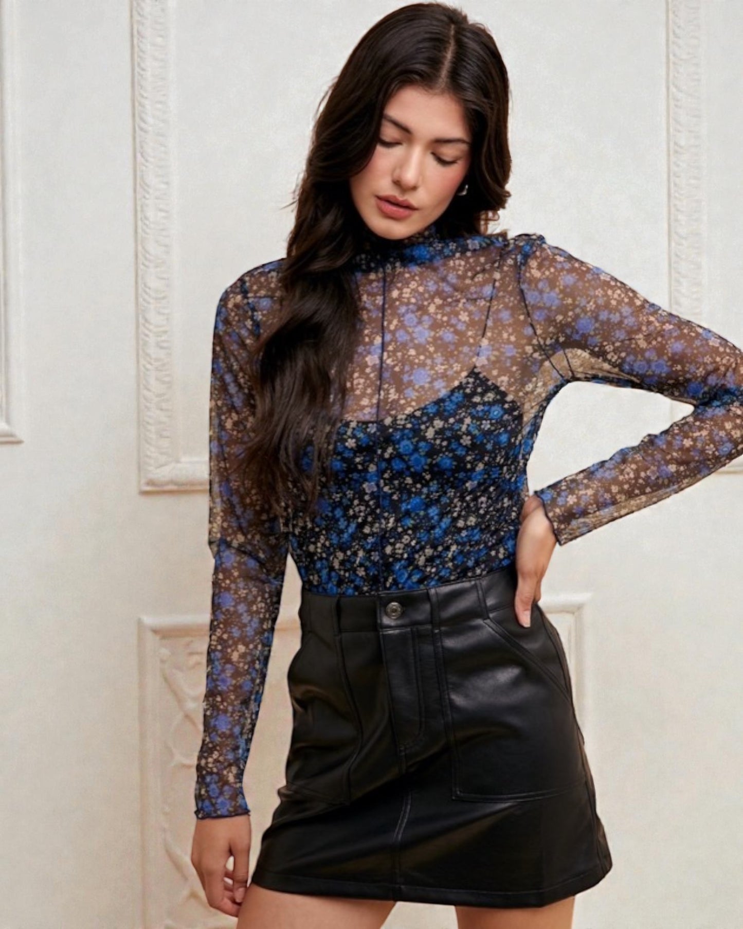 Woman wearing a floral long-sleeve top and black leather skirt against a white wall.