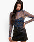 A woman modeling a sheer long-sleeve floral mock-neck top with blue and gold tones over a black underlayer, paired with a black faux-leather mini skirt, against a plain white background.