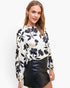 A woman modeling a long-sleeve blouse with a black floral print on an off-white background, paired with a black faux-leather mini skirt, against a plain white studio background.