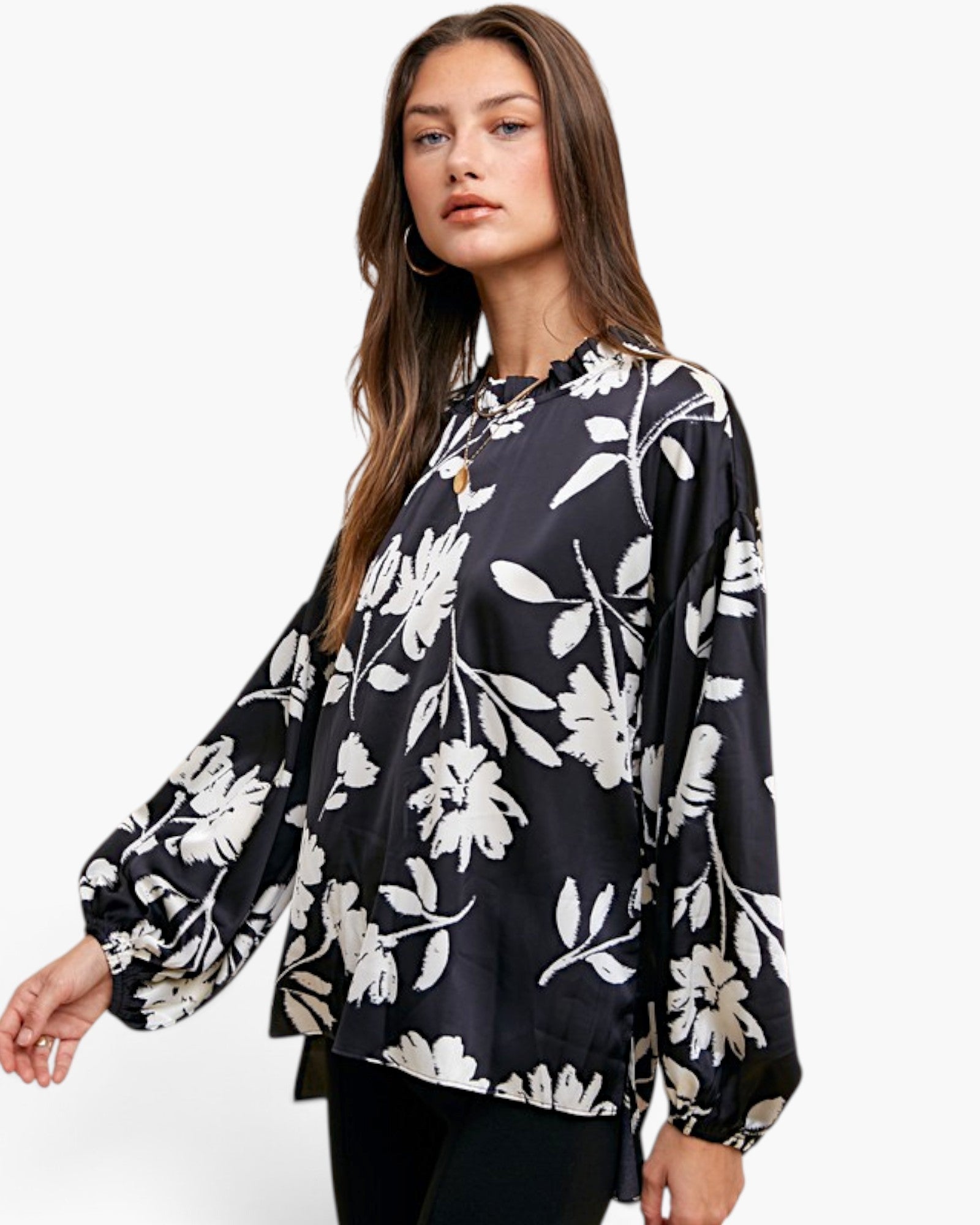 A woman modeling a black long-sleeve blouse with a white floral print, featuring a ruffled high neckline and balloon sleeves, set against a plain white background.