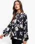 A woman modeling a black long-sleeve blouse with a white floral print, featuring a ruffled high neckline and balloon sleeves, set against a plain white background.