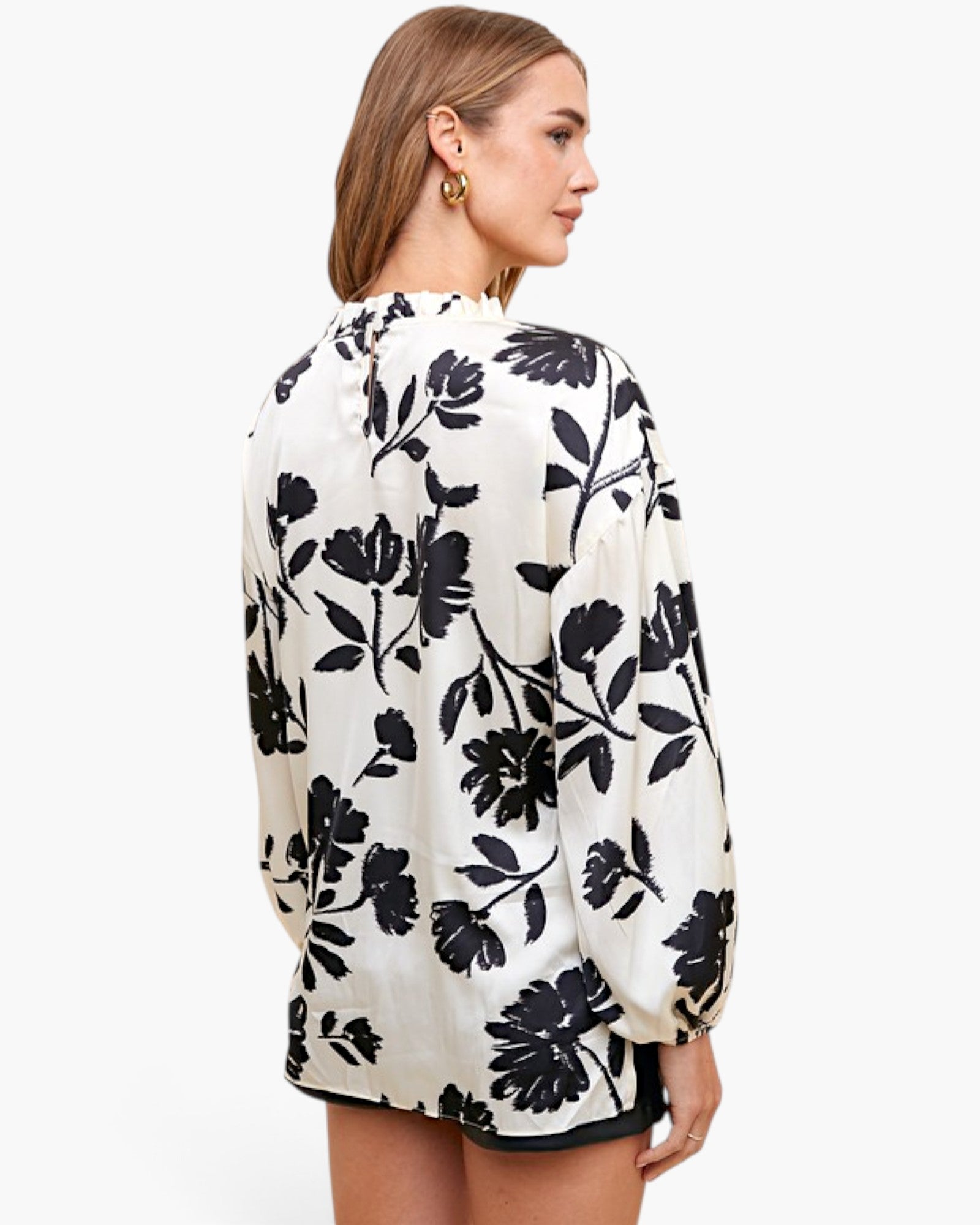 A woman shown from the back wearing a long-sleeve blouse with a black floral print on an off-white background, paired with black shorts, against a plain white studio background.