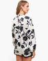 A woman shown from the back wearing a long-sleeve blouse with a black floral print on an off-white background, paired with black shorts, against a plain white studio background.