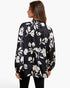 A woman shown from the back wearing a black long-sleeve blouse with a large white floral print, paired with black pants, against a plain white studio background.