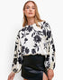A woman wearing a cream-colored blouse with a black floral pattern and long balloon sleeves, paired with a black leather mini skirt, against a plain white studio background.