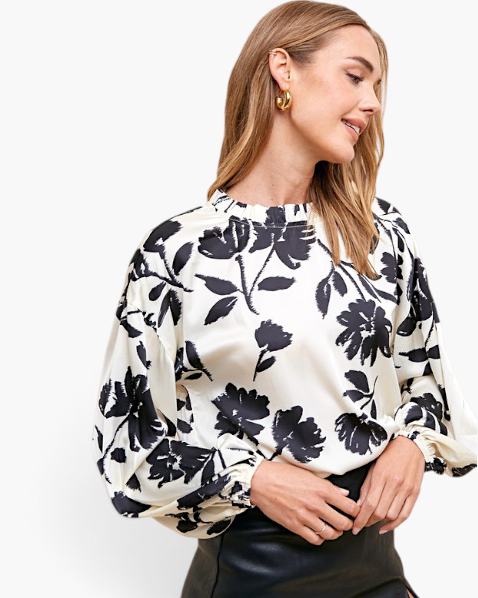 A woman wearing a cream-colored blouse with a bold black floral pattern and balloon sleeves, paired with a black leather skirt, against a plain white studio background.