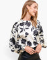 A woman wearing a cream-colored blouse with a bold black floral pattern and balloon sleeves, paired with a black leather skirt, against a plain white studio background.