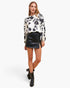 A woman modeling a long-sleeve blouse with a black floral print on a cream background, paired with a black faux-leather mini skirt and black ankle boots, against a light gray studio background.