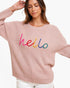 A woman wearing a light pink ribbed knit sweater with the word 'hello' embroidered across the chest in bright multicolored letters, paired with white pants against a plain white background.