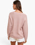 A woman shown from the back wearing a dusty pink ribbed knit sweater and white frayed denim shorts against a plain white studio background.