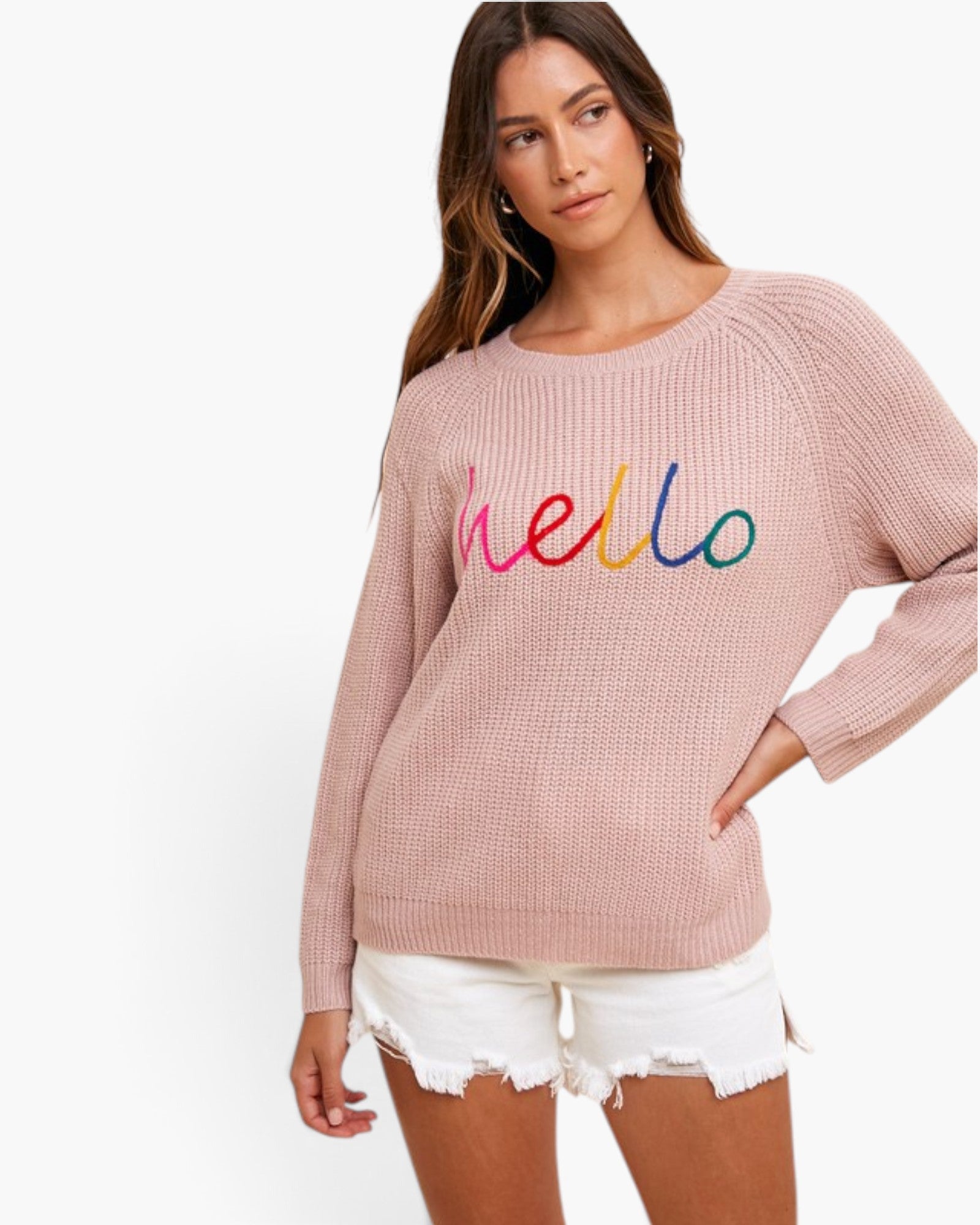 A woman wearing a dusty pink waffle-knit sweater with 'hello' embroidered in colorful script across the chest, paired with white frayed denim shorts, against a plain white background.