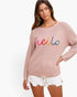 A woman wearing a dusty pink waffle-knit sweater with 'hello' embroidered in colorful script across the chest, paired with white frayed denim shorts, against a plain white background.