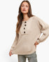 A woman wearing a beige waffle-knit sweater with a buttoned split neckline and drawstring detail, paired with gray jeans, against a plain white studio background.