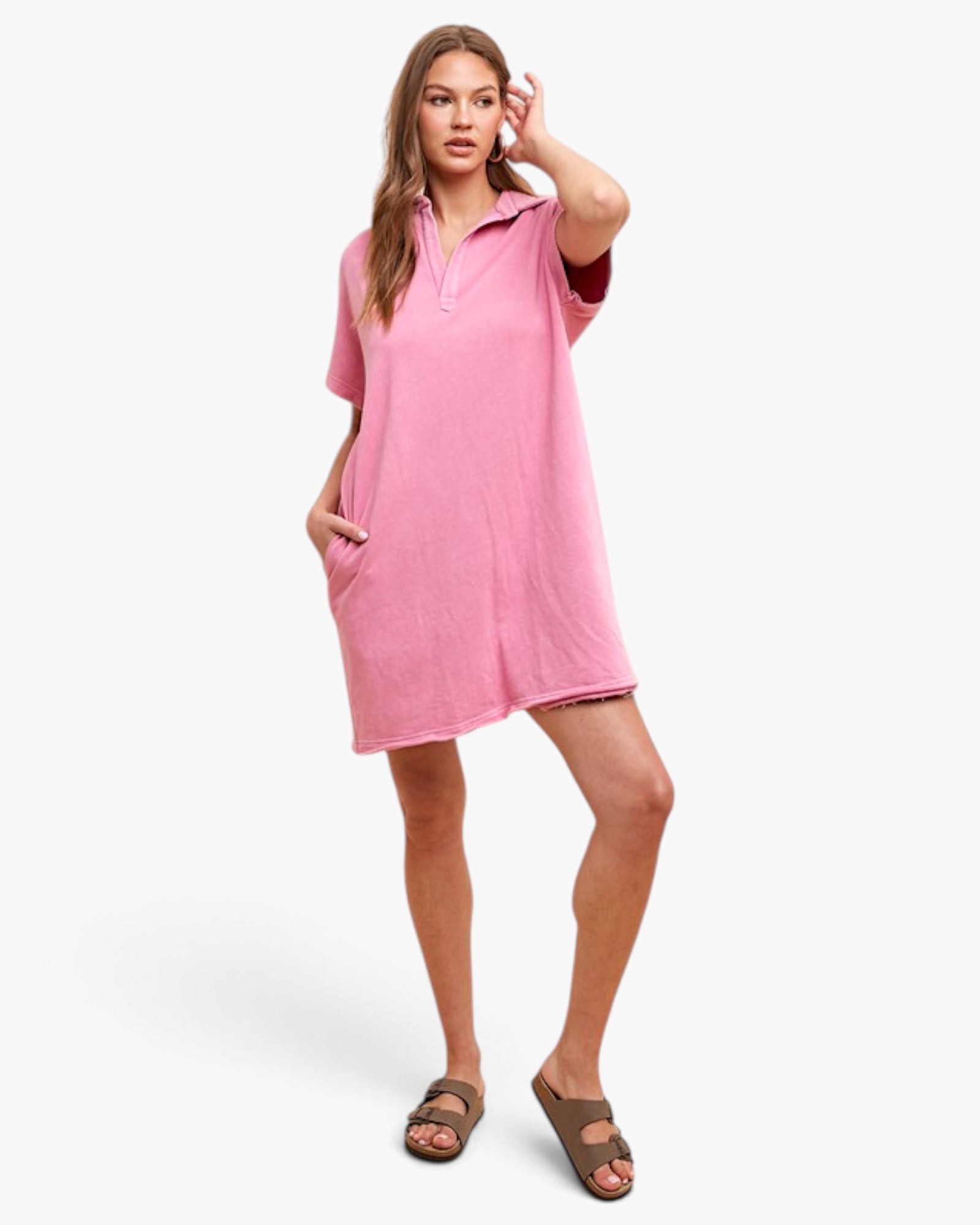 A woman modeling a pink short-sleeve polo shirt dress with side pockets, paired with brown slide sandals, against a light gray studio background.
