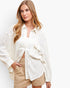 A woman wearing a textured off-white button-up shirt with ruffle details and beige shorts, photographed against a clean white studio background.