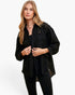 A woman modeling a black textured, semi-sheer button-up shirt with long sleeves and a high-low hem, paired with black pants against a plain white studio background.