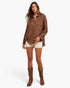 A woman modeling a brown textured button-up shirt with cream denim shorts and brown leather cowboy boots, photographed against a clean white studio background.