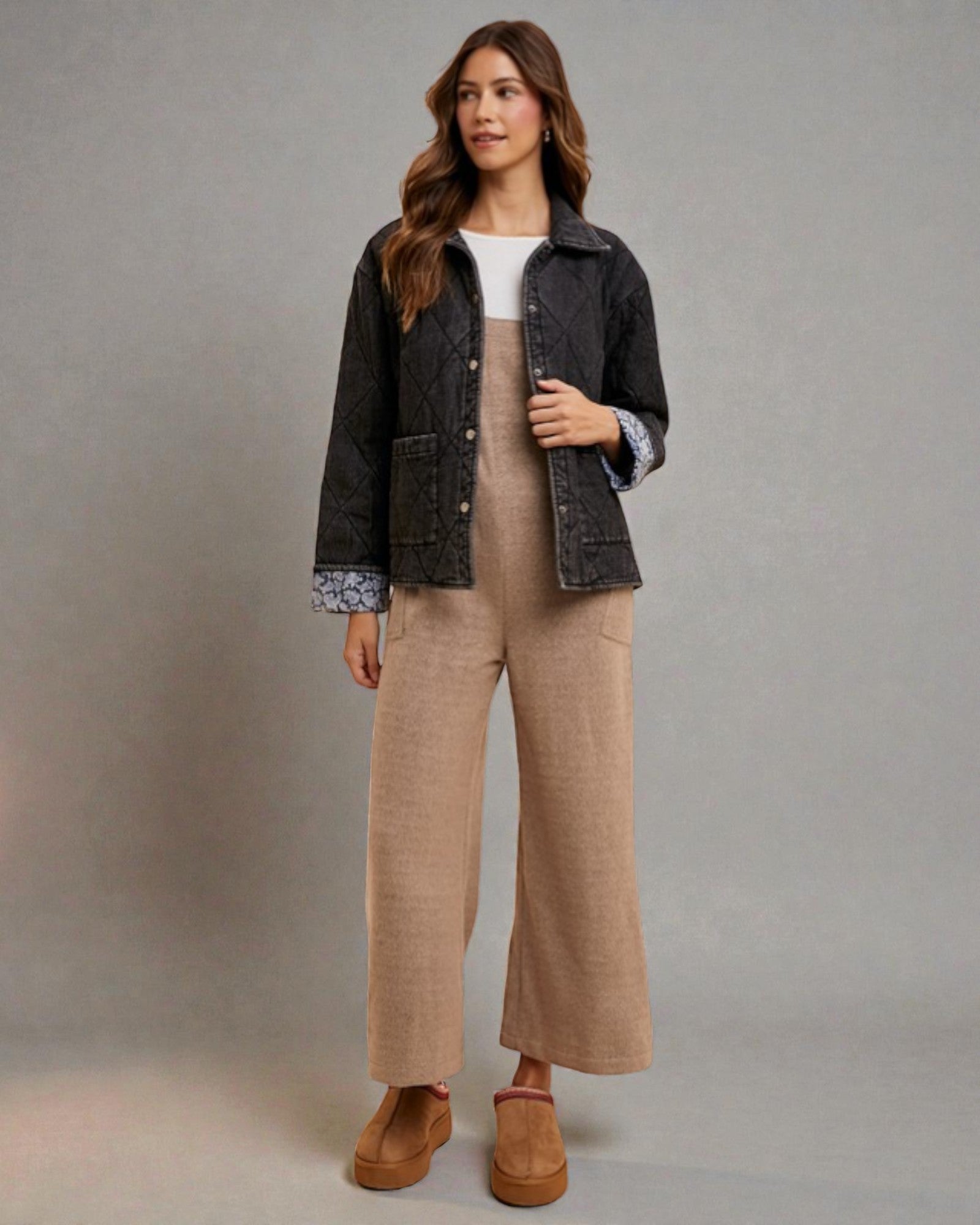 Woman wearing a black jacket, white shirt, beige pants, and brown shoes against a gray background