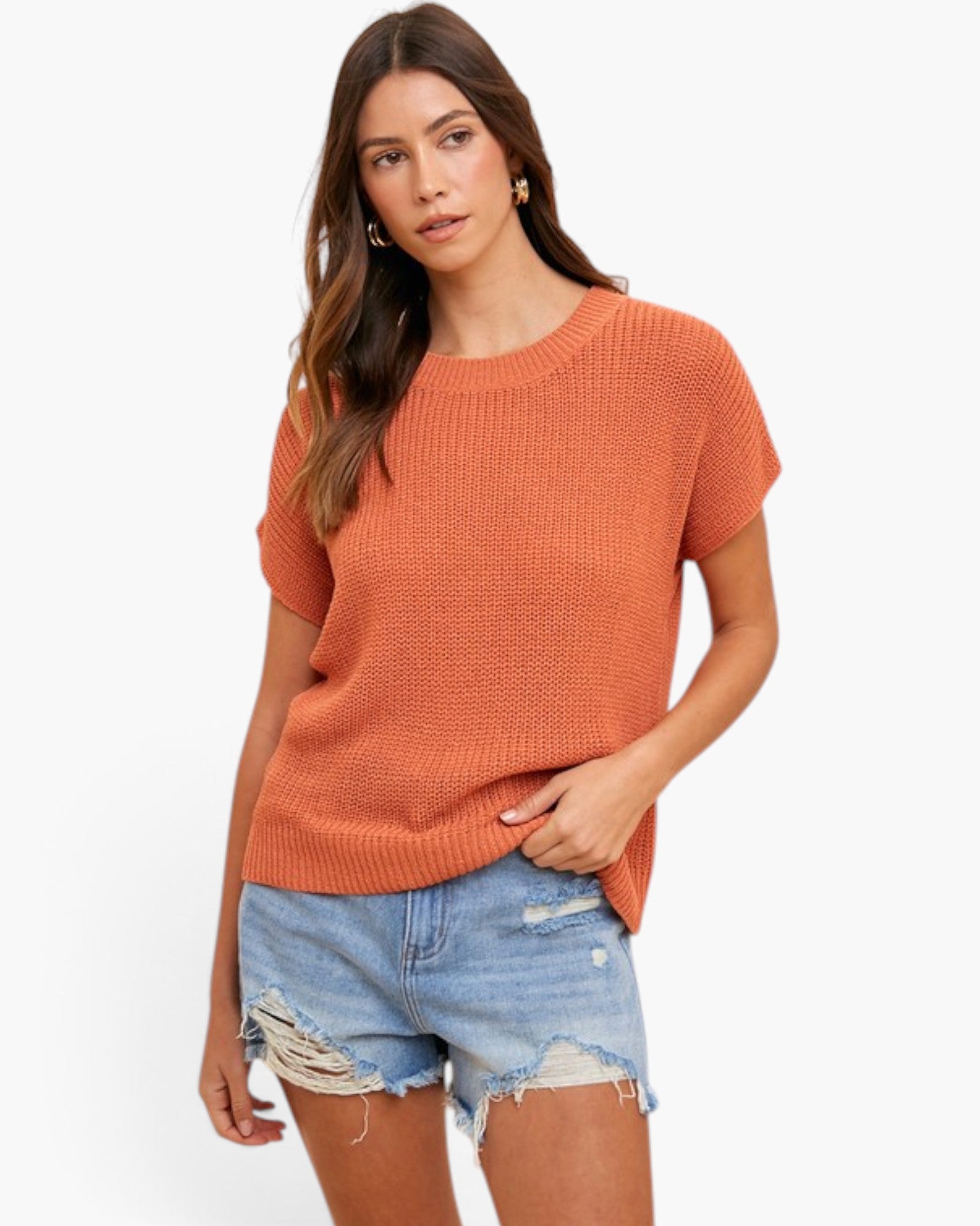 A woman modeling a short-sleeve, crew-neck knit sweater in a warm rust orange color, paired with distressed denim shorts, against a plain white studio background.
