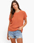 A woman modeling a short-sleeve, crew-neck knit sweater in a warm rust orange color, paired with distressed denim shorts, against a plain white studio background.