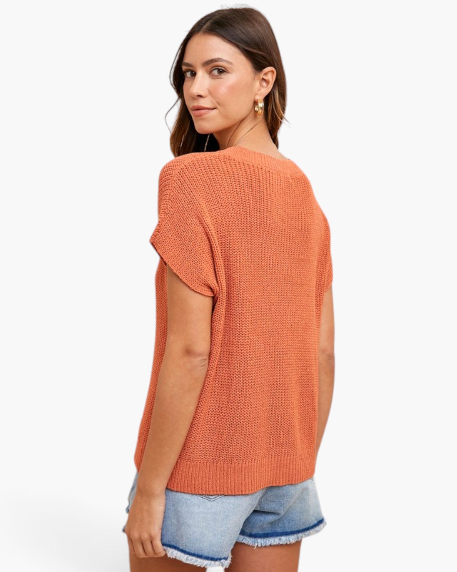 A woman wearing a textured, short-sleeve knit top in a muted orange/coral color, paired with denim shorts, shown from a three-quarters back view against a plain white background.