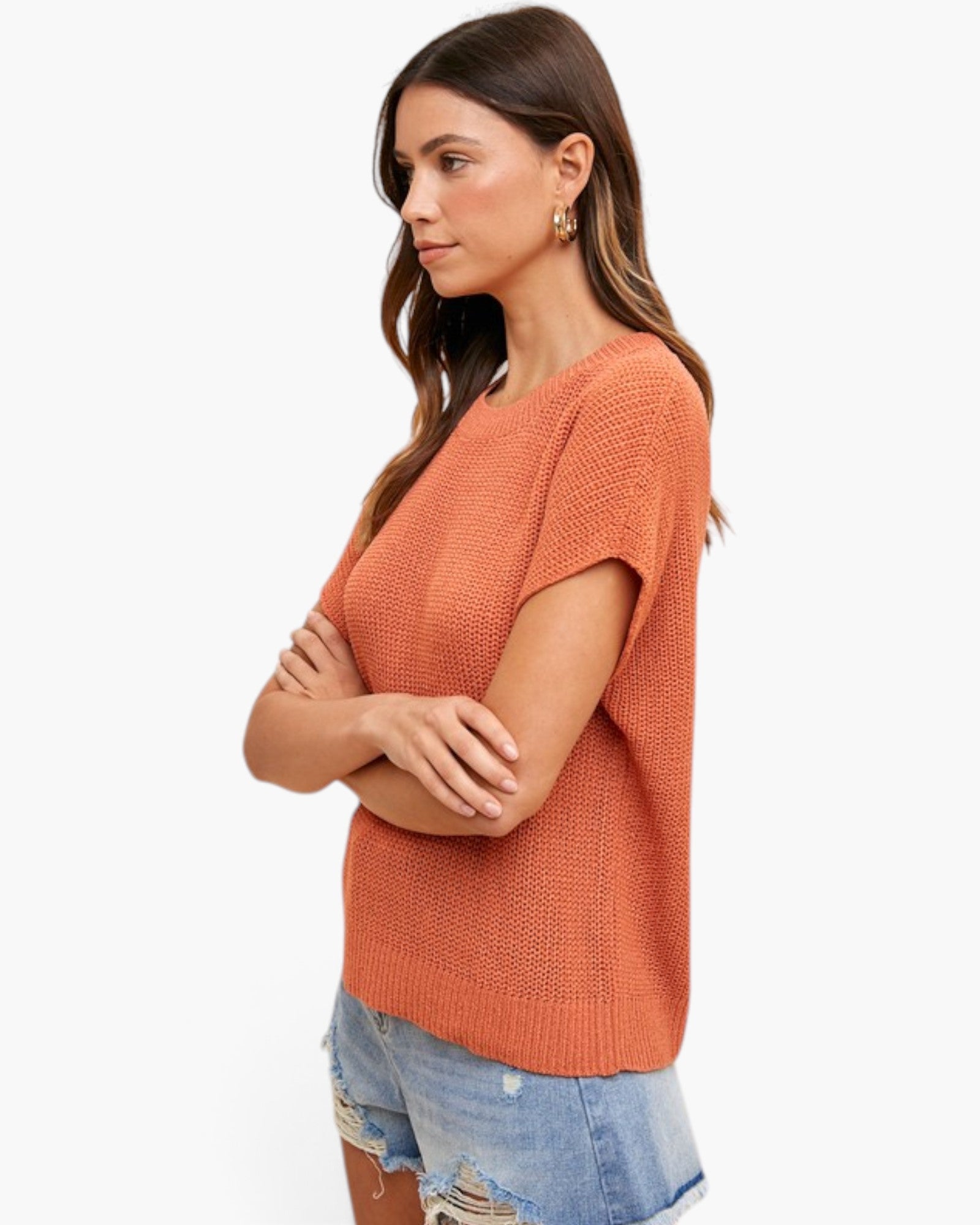 A woman wearing a textured short-sleeve knit top in a warm rust orange color, paired with light-wash denim shorts, photographed against a clean white background.