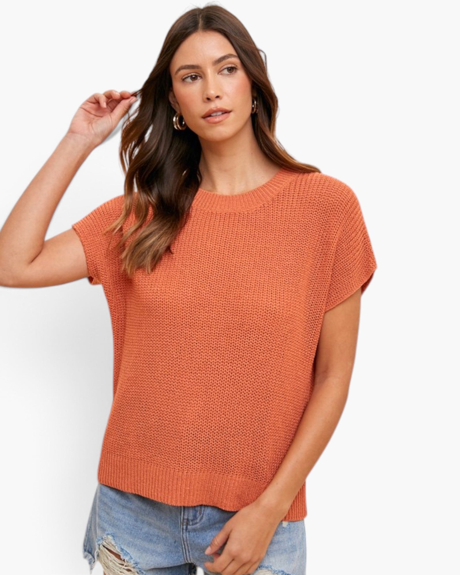 A woman modeling a short-sleeve, textured knit crewneck sweater in a warm rust-orange color, paired with distressed blue jeans against a clean white background.