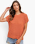 A woman modeling a short-sleeve, textured knit crewneck sweater in a warm rust-orange color, paired with distressed blue jeans against a clean white background.
