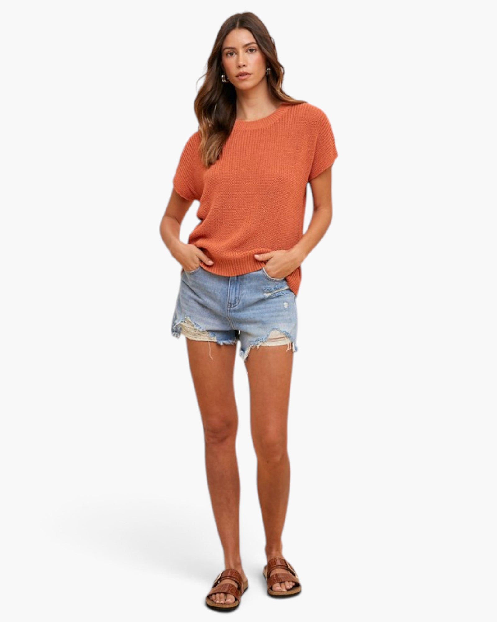 A woman modeling a short-sleeve ribbed knit top in a muted burnt orange color, paired with light-wash distressed denim shorts and brown leather slide sandals, against a plain white studio background.