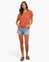 A woman modeling a short-sleeve ribbed knit top in a muted burnt orange color, paired with light-wash distressed denim shorts and brown leather slide sandals, against a plain white studio background.
