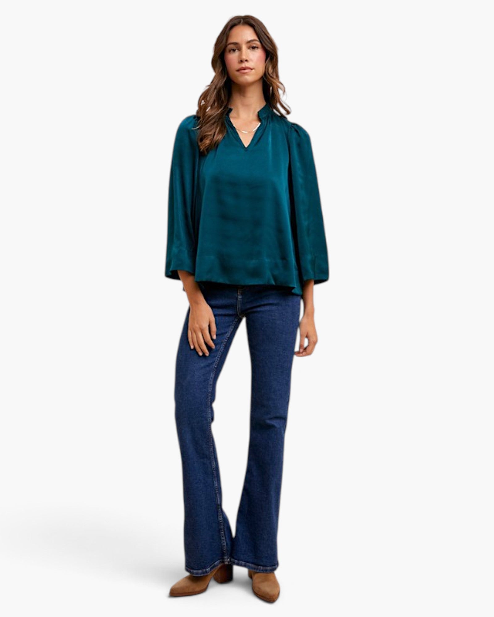 A woman modeling a teal green satin long-sleeve blouse with a split V-neckline, paired with dark blue flared jeans and brown ankle boots, against a plain white studio background.