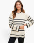A woman modeling a cream-colored knit sweater with horizontal black stripes, paired with black pants, against a plain white studio background.
