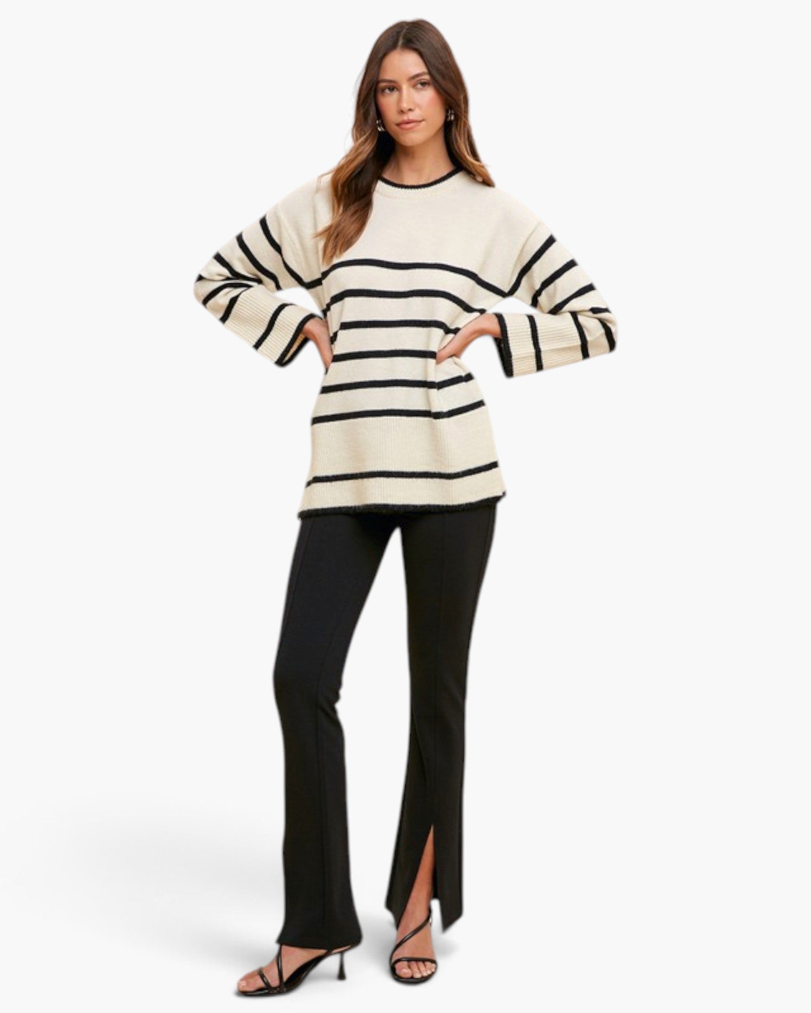 A woman modeling a cream knit sweater with black horizontal stripes and black flared pants with front slits, paired with black heeled sandals, against a plain white studio background.