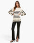 A woman modeling a cream knit sweater with black horizontal stripes and black flared pants with front slits, paired with black heeled sandals, against a plain white studio background.