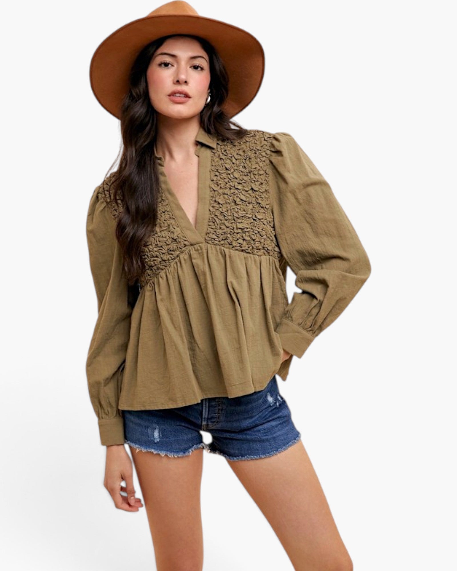 A woman modeling a long-sleeve olive green blouse with a lace-trimmed V-neck and peplum waist, paired with denim shorts and a wide-brim tan hat against a light gray studio background.