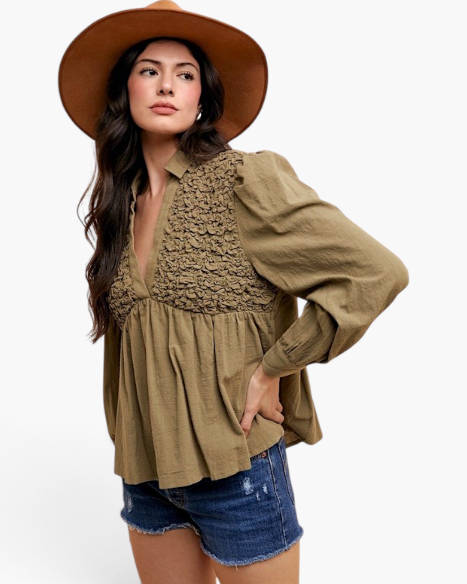 A woman wearing a brown wide-brim hat, an olive green long-sleeve blouse with a textured lace-like yoke, and blue denim shorts, posed against a plain light gray studio background.