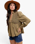 A woman wearing a brown wide-brim hat, an olive green long-sleeve blouse with a textured lace-like yoke, and blue denim shorts, posed against a plain light gray studio background.