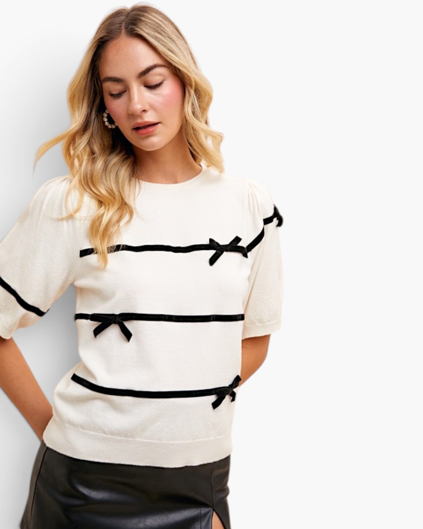 A woman wearing a cream-colored short-sleeve knit top with black horizontal trim and bow details, paired with black leather pants, against a plain white studio background.