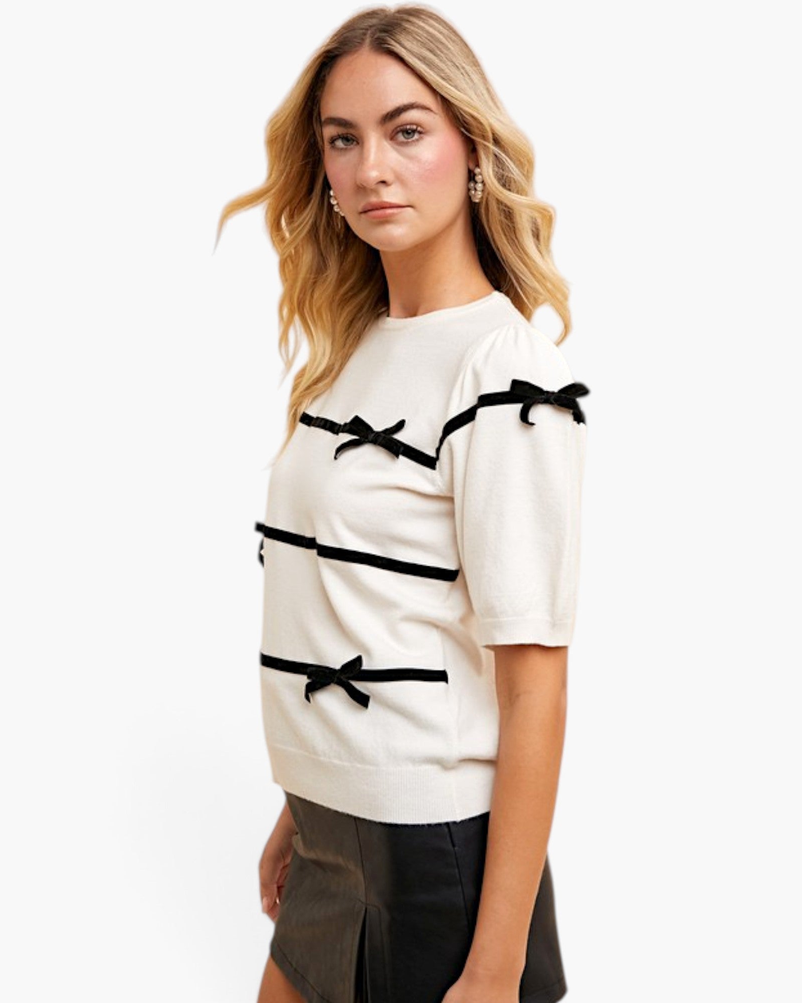 A woman modeling a cream-colored short-sleeve knit top with black ribbon trim and bow details, paired with a dark skirt, against a plain white studio background.