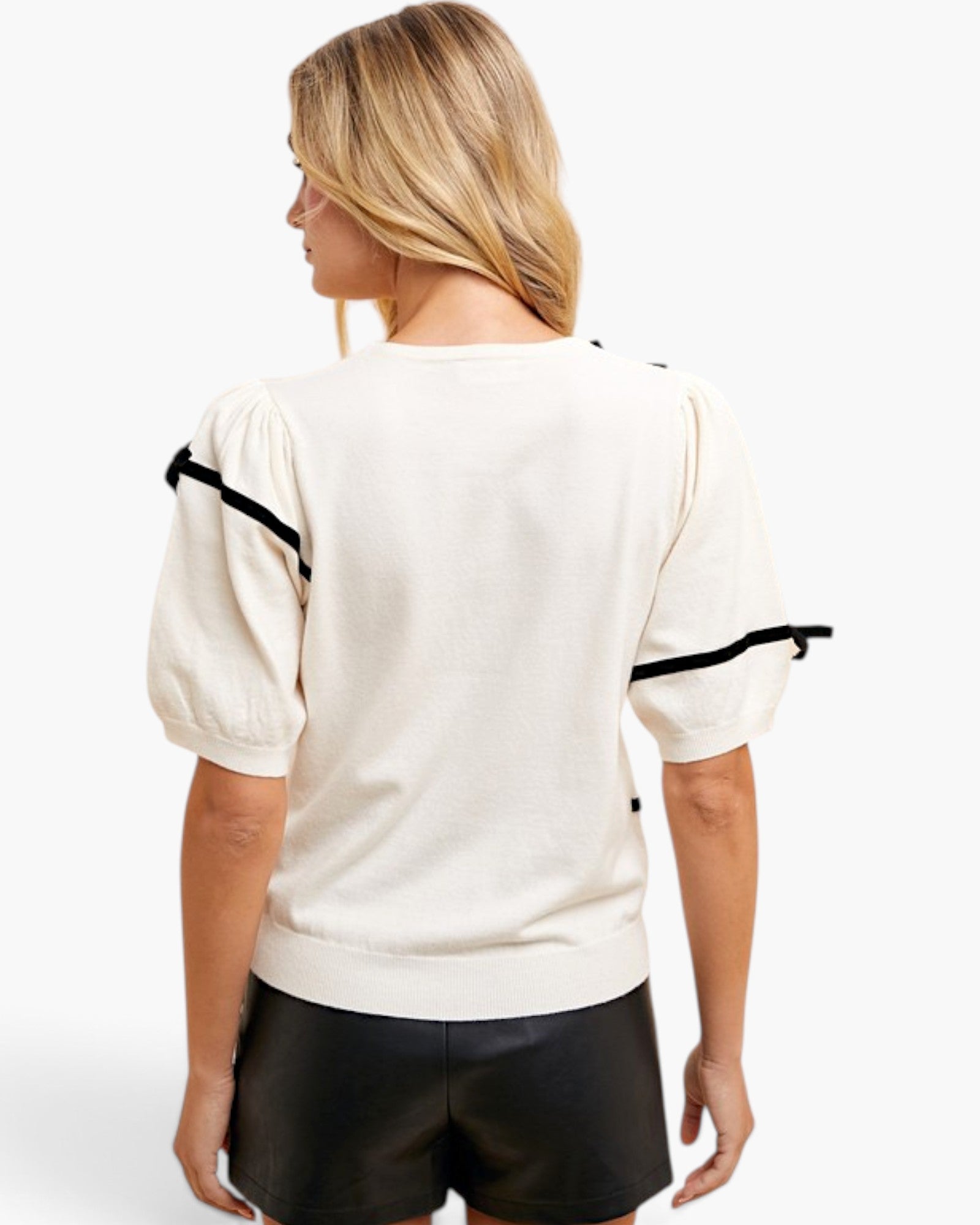 A woman shown from the back wearing a cream-colored knit top with black ribbon accents on the puff sleeves, paired with black shorts, against a plain white background.