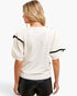 A woman shown from the back wearing a cream-colored knit top with black ribbon accents on the puff sleeves, paired with black shorts, against a plain white background.
