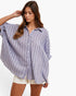 A woman wearing a loose, oversized button-up shirt with vertical blue and white stripes, paired with cream shorts, against a plain white studio background.