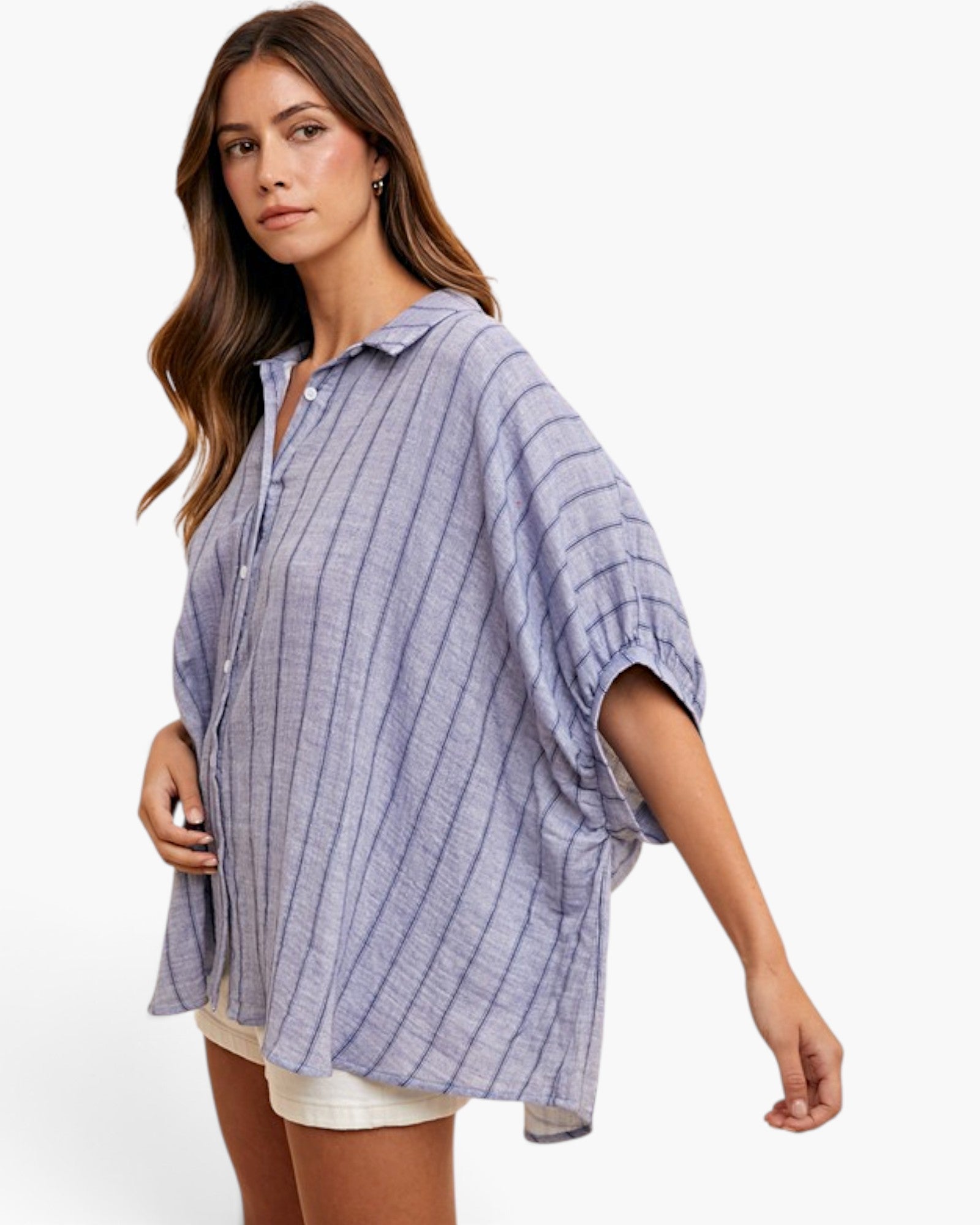 A woman wearing a loose, oversized button-up shirt with vertical blue and white stripes, styled with white shorts against a clean white background.