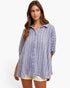 A woman modeling a loose, short-sleeve button-up shirt with a blue and white vertical stripe pattern, paired with white shorts, against a plain white studio background.